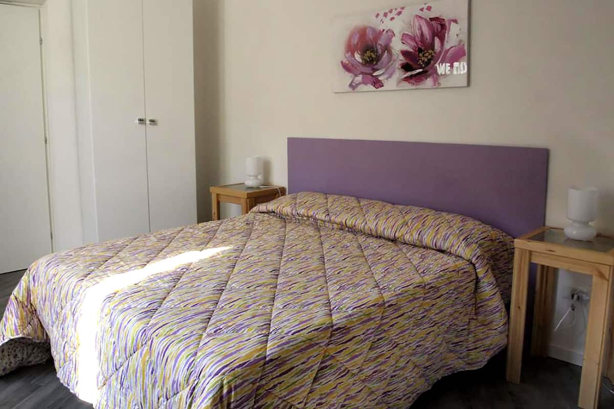 A bedroom with a bed, a nightstand, a wall-mounted lamp, and a door.