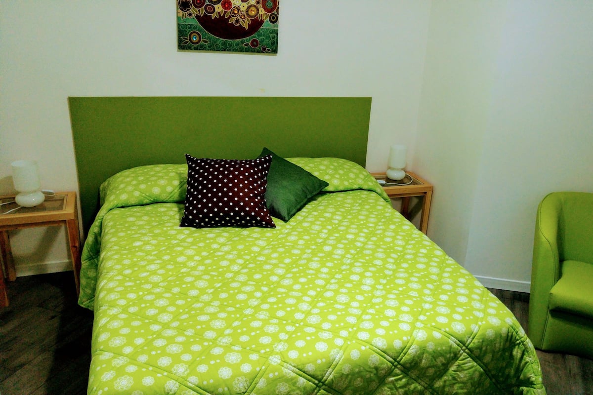 A room with a bed, a green chair, and a nightstand with a lamp.