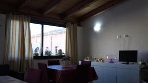 Comedor con una mesa dispuesta para cuatro, zona de cocina con televisión y armarios, y una ventana con cortinas.