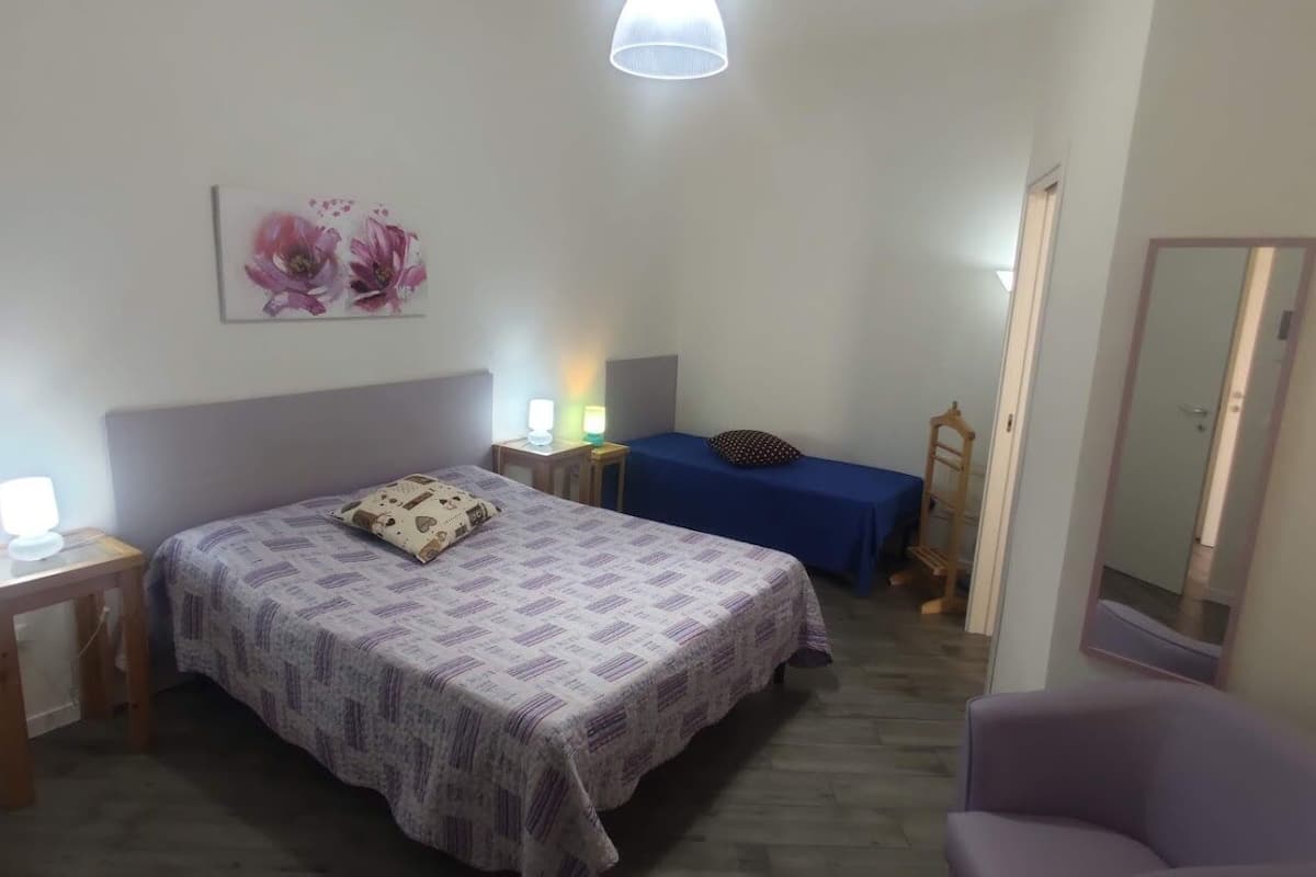 A bedroom with a bed, a bedside table with a lamp, a chair, a small table with a lamp, and a mirror on the wall.