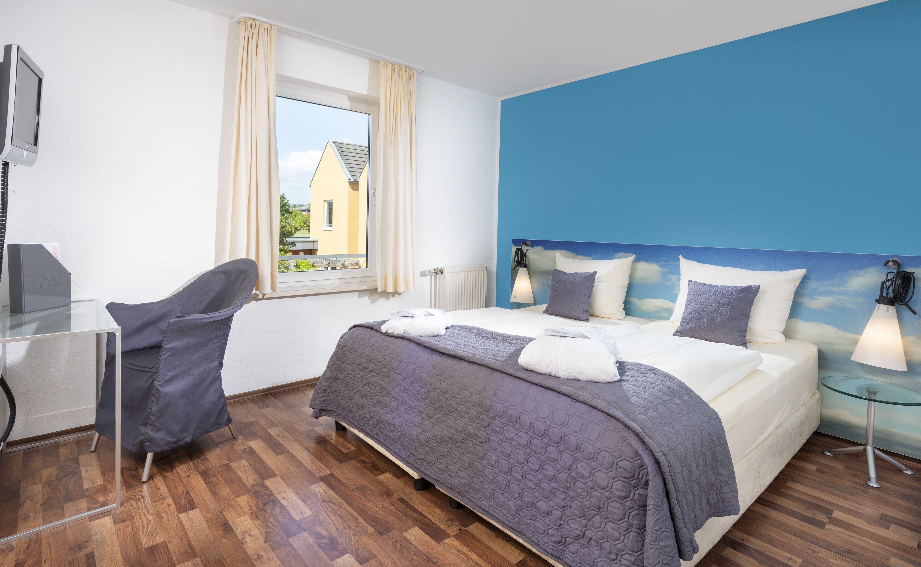 Une chambre d’hôtel avec un lit, un bureau, une chaise, une fenêtre avec des rideaux et une vue sur un bâtiment à l’extérieur.