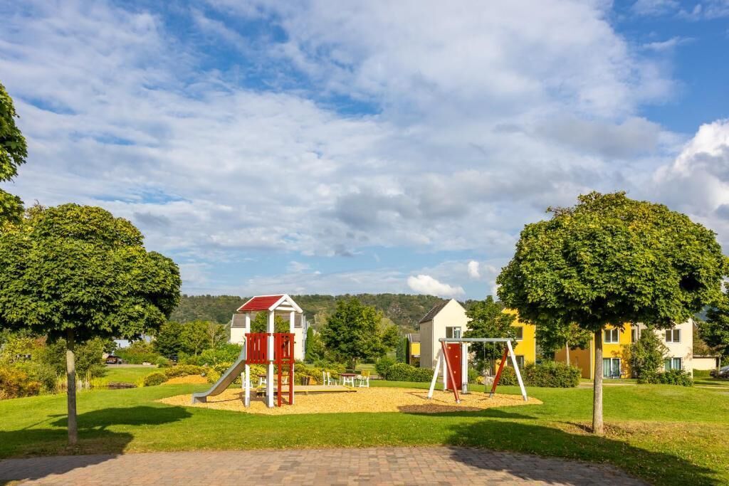 A playground with red and white equipment, surrounded by green grass and trees, with houses in the background.