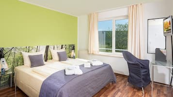 Quarto de hotel com cama, escrivaninha, cadeira, janela com cortinas e vista para árvores.