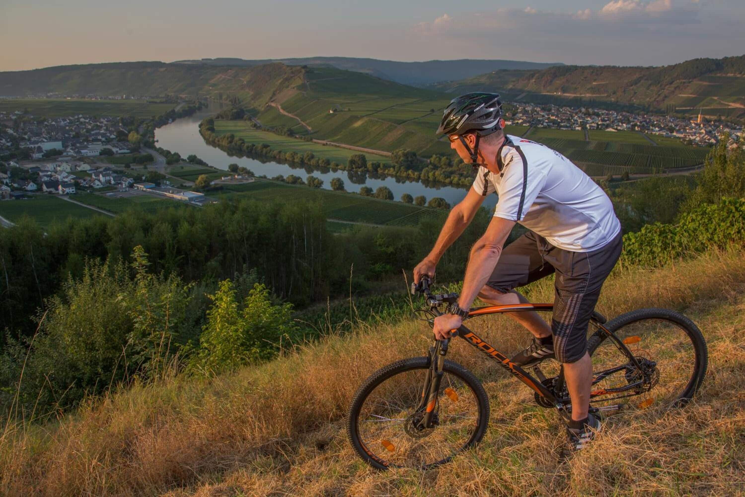 A cyclist on a mountain bike trail with a scenic view of a river, town, and hills in the background.