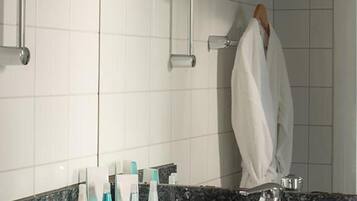 Et badeværelse med en hvid flisevæg, en vask med en granitbordplade og et spejl. Der er toiletartikler på bordet og en håndklædestang med håndklæder.