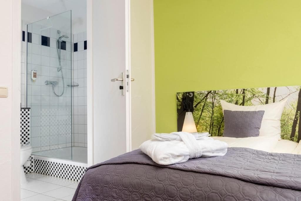 A hotel room with a glass-enclosed shower, a bed with a grey quilt, and a view of trees through a window.