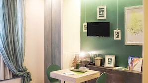 A small, cozy room with a green wall, a flat-screen TV, a desk with a chair, and a window with curtains. - Spazio Bianco (Ivrea)