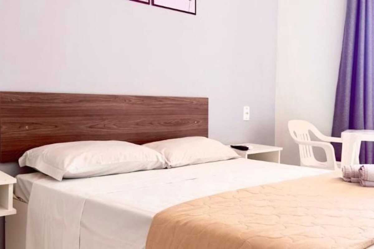 Quarto de hotel com cabeceira de madeira, cama com roupa de cama branca, mesa de cabeceira, uma cadeira e janela com cortinas roxas.