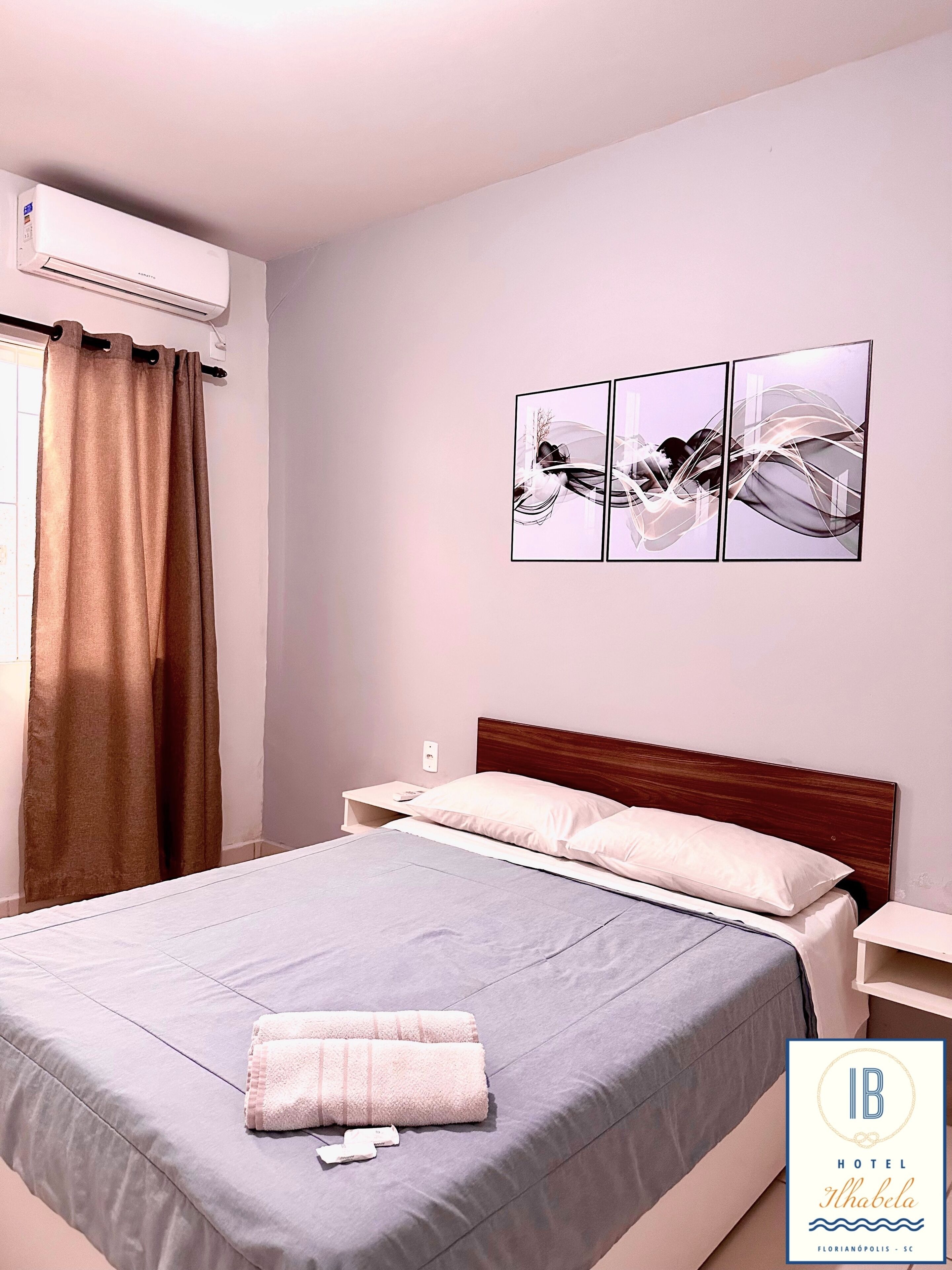 Une chambre d’hôtel comprenant un lit, une table de chevet, un appareil de climatisation, une fenêtre avec des rideaux et une œuvre d’art accrochée au mur.