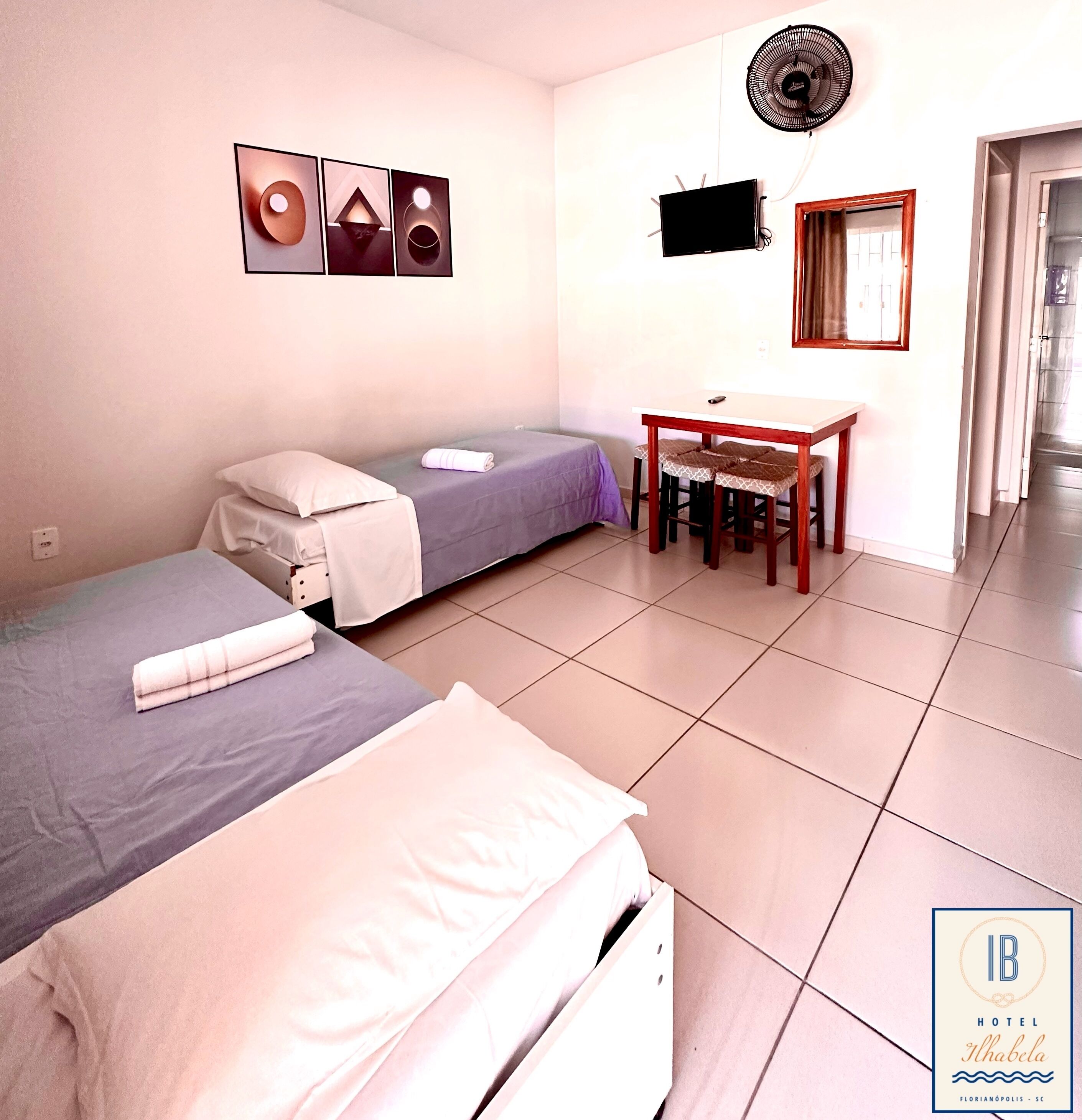 A hotel room with two beds, a small table with chairs, a TV, and a fan.