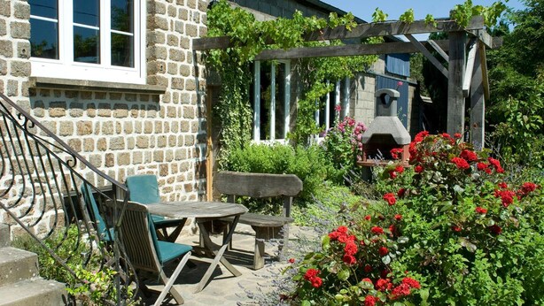 Un bâtiment en pierre doté d’une pergola en bois, d’un espace extérieur aménagé pour s’asseoir et d’un jardin orné de fleurs rouges.