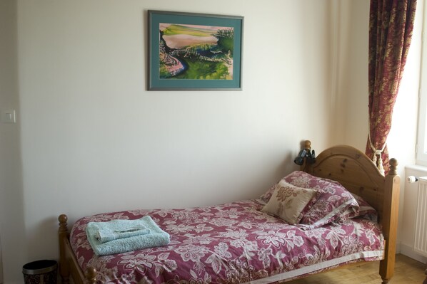 A single bed with floral bedding, a blue towel, and a framed painting on the wall. - La Neslière – Chambres d’hôtes Bio (Buais-les-Monts)