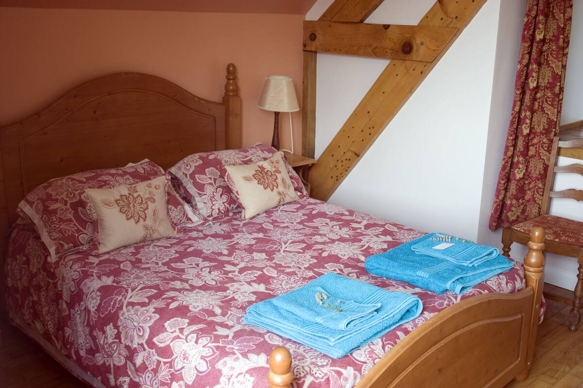 Un lit avec une literie à motifs floraux, une tête de lit en bois et une table de chevet avec une lampe. Des serviettes bleues sont pliées sur le lit.