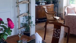 Un coin repas avec une table en bois dressée pour deux personnes, entourée de chaises en bois. On y voit une échelle ornée de plantes grimpantes, un tableau accroché au mur et, au fond, des plantes en pot.
