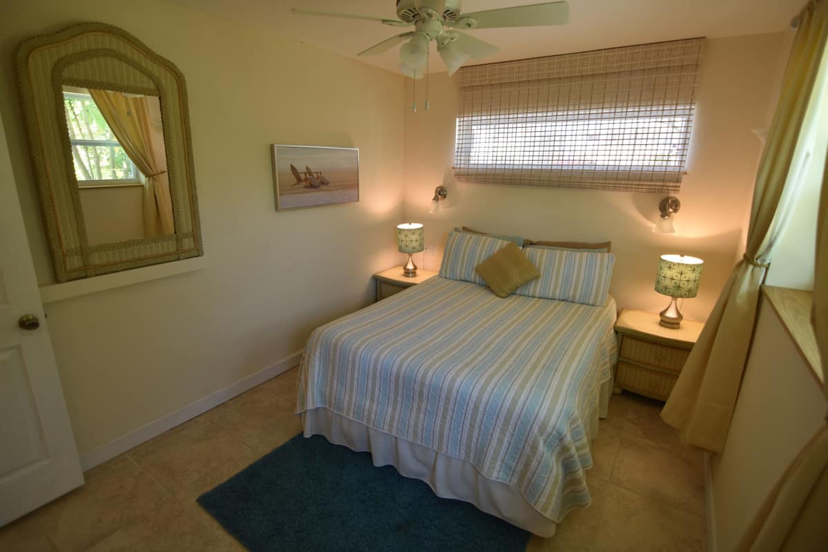 A bedroom with a bed, two bedside tables with lamps, a ceiling fan, a window with blinds, a mirror, and a door.