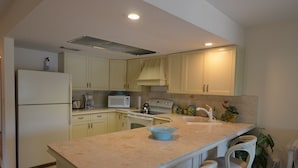 A kitchen with light yellow cabinets, a white refrigerator, a microwave, a stove, and a sink. There are two white wicker chairs at the island.