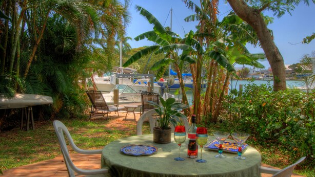 An outdoor dining area with a table set for two, surrounded by chairs, a potted plant, and a view of the marina with boats.