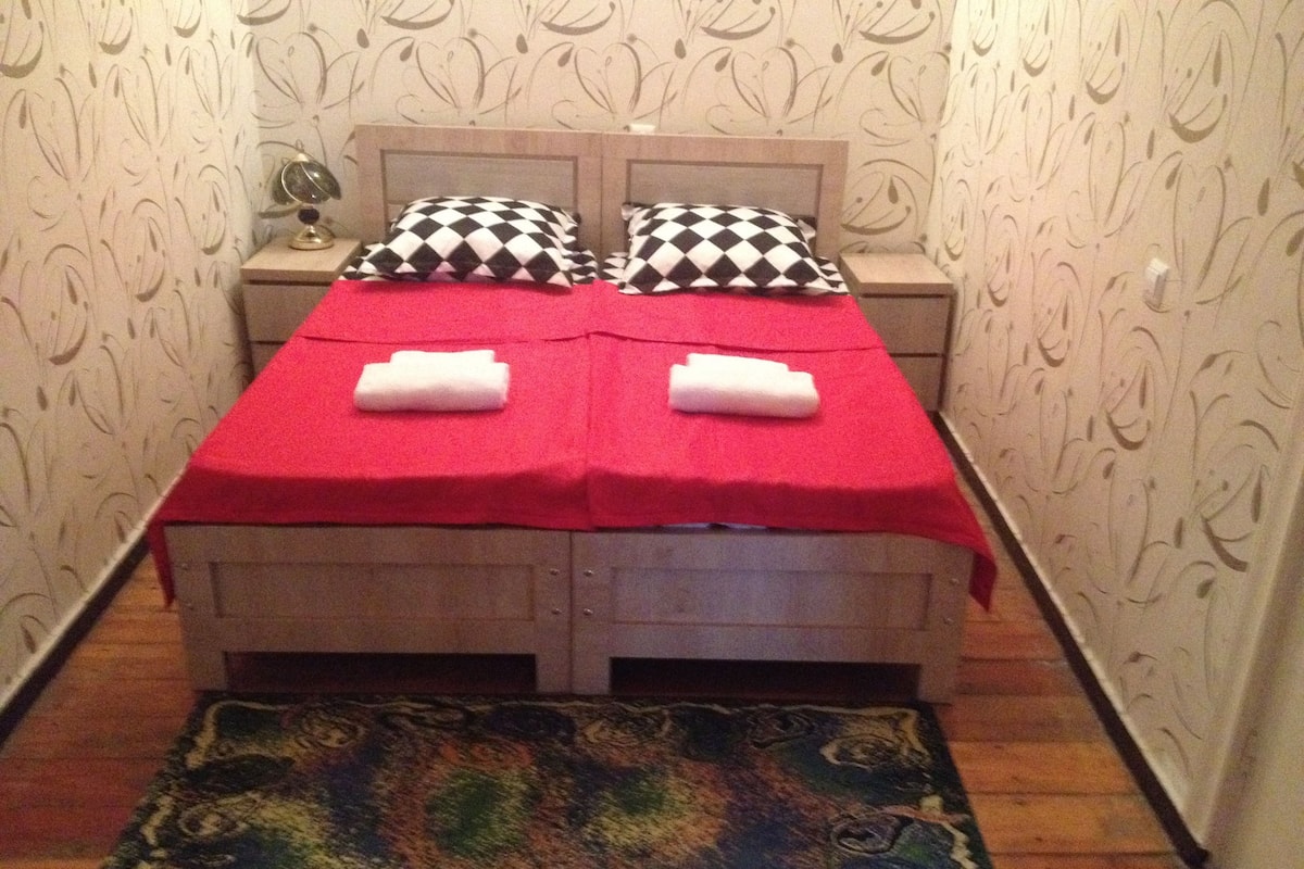 A small, neatly made bed with a red blanket and two pillows, a wooden headboard, a nightstand with a lamp, and a colorful rug on the floor.