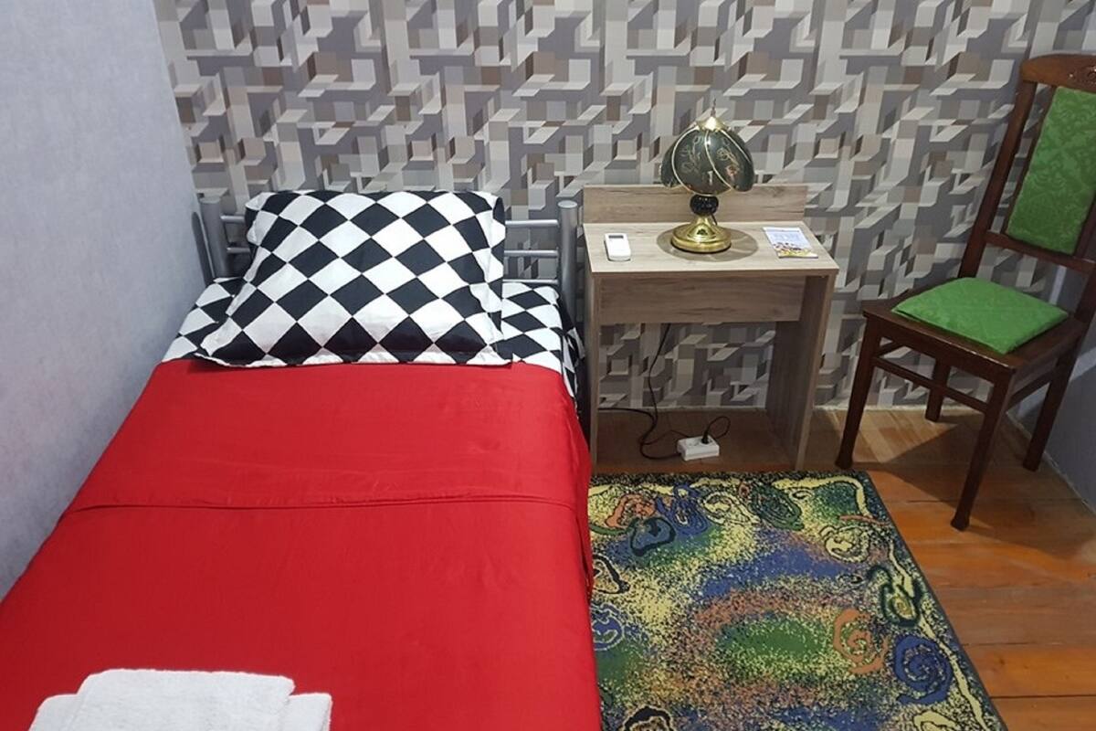 A small, neatly arranged room with a single bed, a chair, a nightstand with a lamp, and a patterned rug.