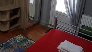 Quarto com guarda-roupa de madeira, cama com colcha vermelha, janela com cortinas leves e radiador.