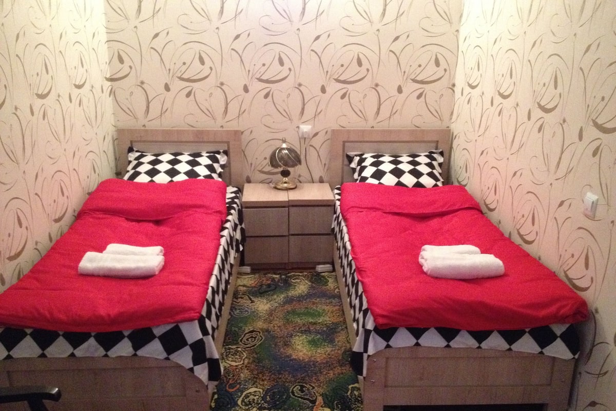 A room with two single beds, each with a red blanket and a checkered pillow. There is a small table between the beds with a lamp on it. The walls have a floral wallpaper pattern.