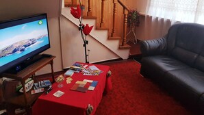 Uma sala de estar com televisão, uma mesa vermelha com revistas e cartas, um sofá de couro preto e uma escada com grades de madeira.