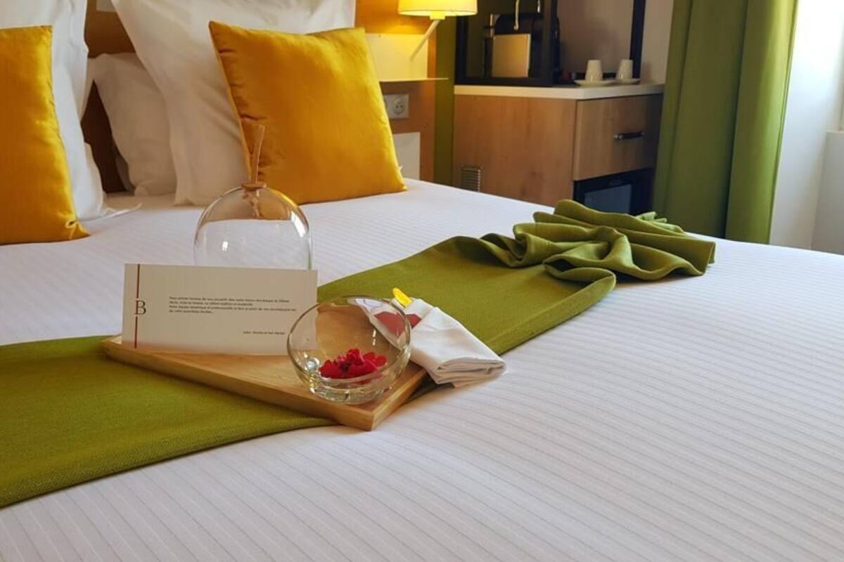 A neatly made hotel bed with a green towel, a glass jar with flowers, and a card on a wooden tray.