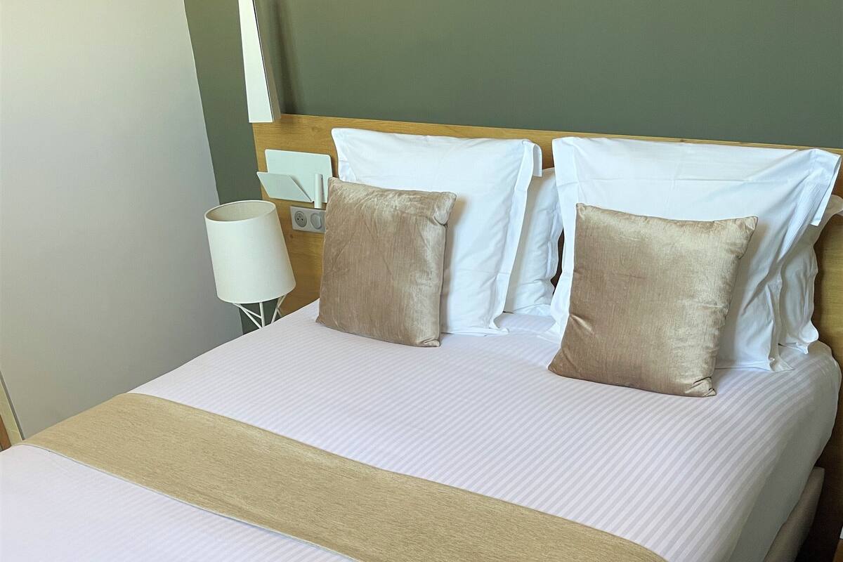 A neatly made bed with a striped bedspread and two beige pillows, a bedside table with a lamp, and a floral-patterned carpet.