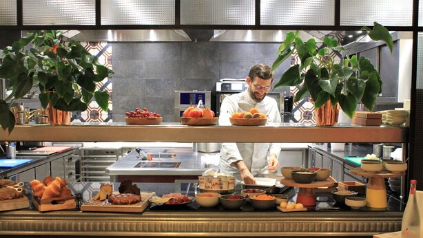 Uno chef prepara dei cibi in una cucina moderna, con diversi ingredienti e piatti esposti.