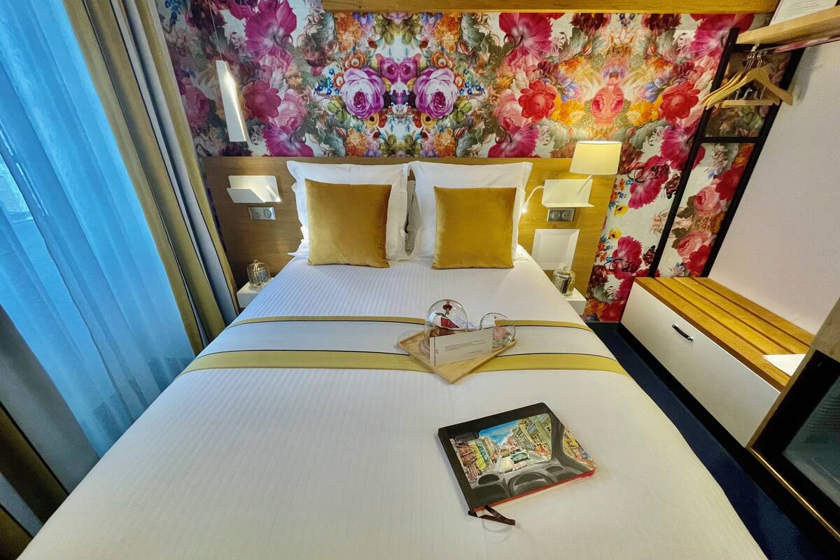 A hotel room with a bed, a bedside table, a nightstand, a lamp, and a floral wallpaper.