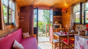 Un acogedor interior de cabaña con techo de madera, un sofá rojo con cojines estampados, una mesa de comedor con sillas y vistas a la vegetación exterior.