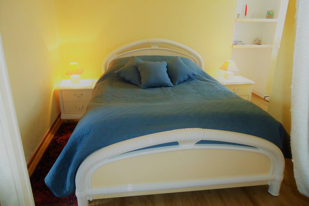 Een klein, verzorgd bed met een wit frame en blauw beddengoed, een witte nachtkastje met een lamp, en een klein plankje met spullen erop.