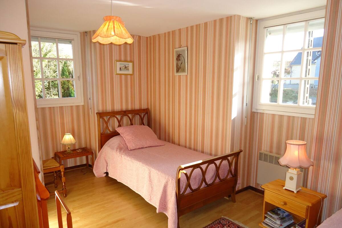 Een slaapkamer met een houten bed, gestreept behang, een raam, een nachtkastje met een lamp en een vloerkleed.