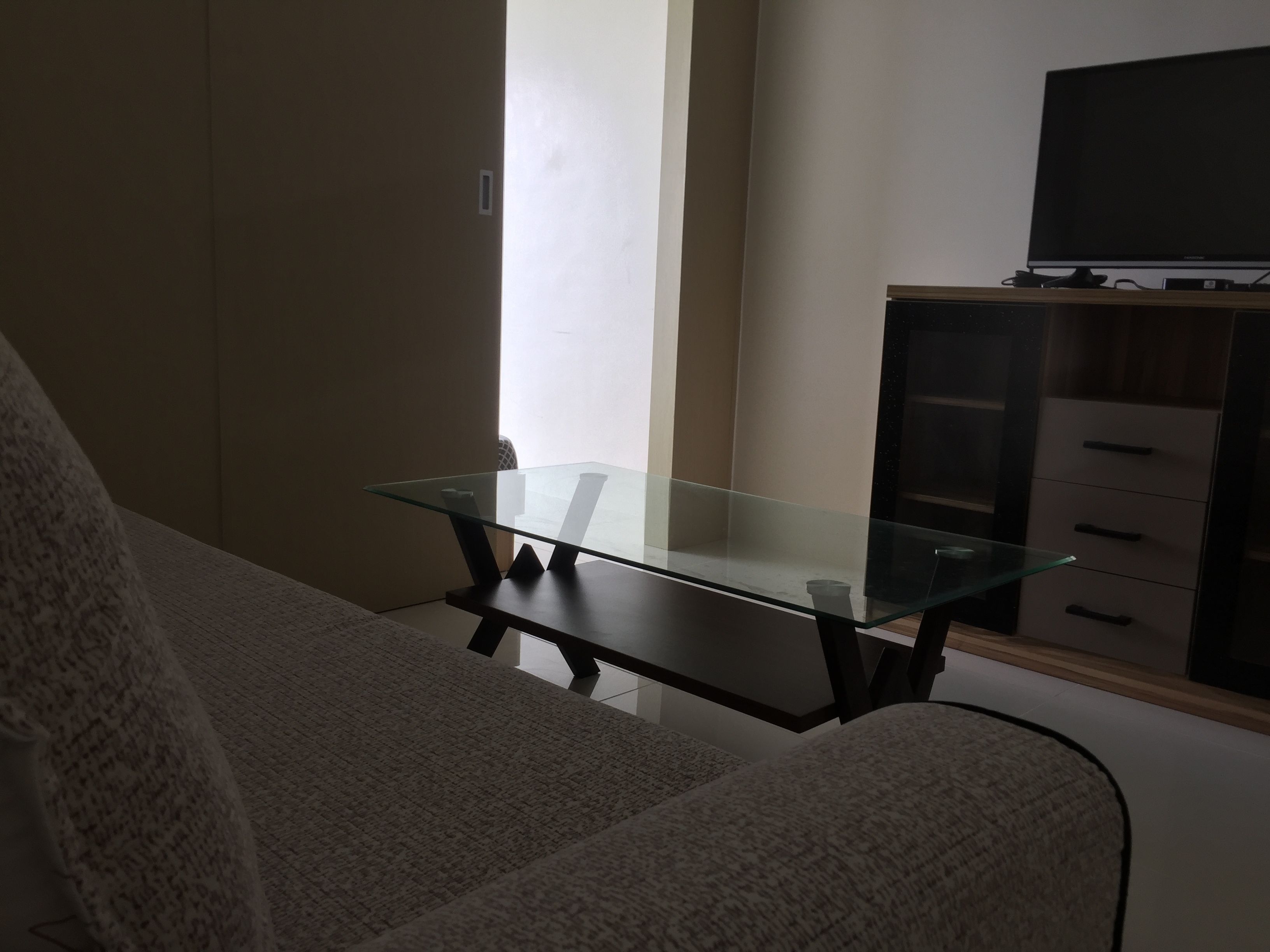 Una sala de estar con una mesa de centro de vidrio, un sofá, un televisor sobre un mueble bajo y un armario con cajones.