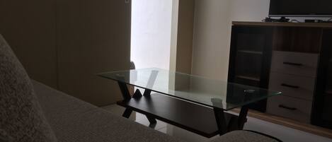 A living room with a glass-top coffee table, a sofa, a TV on a stand, and a cabinet with drawers.