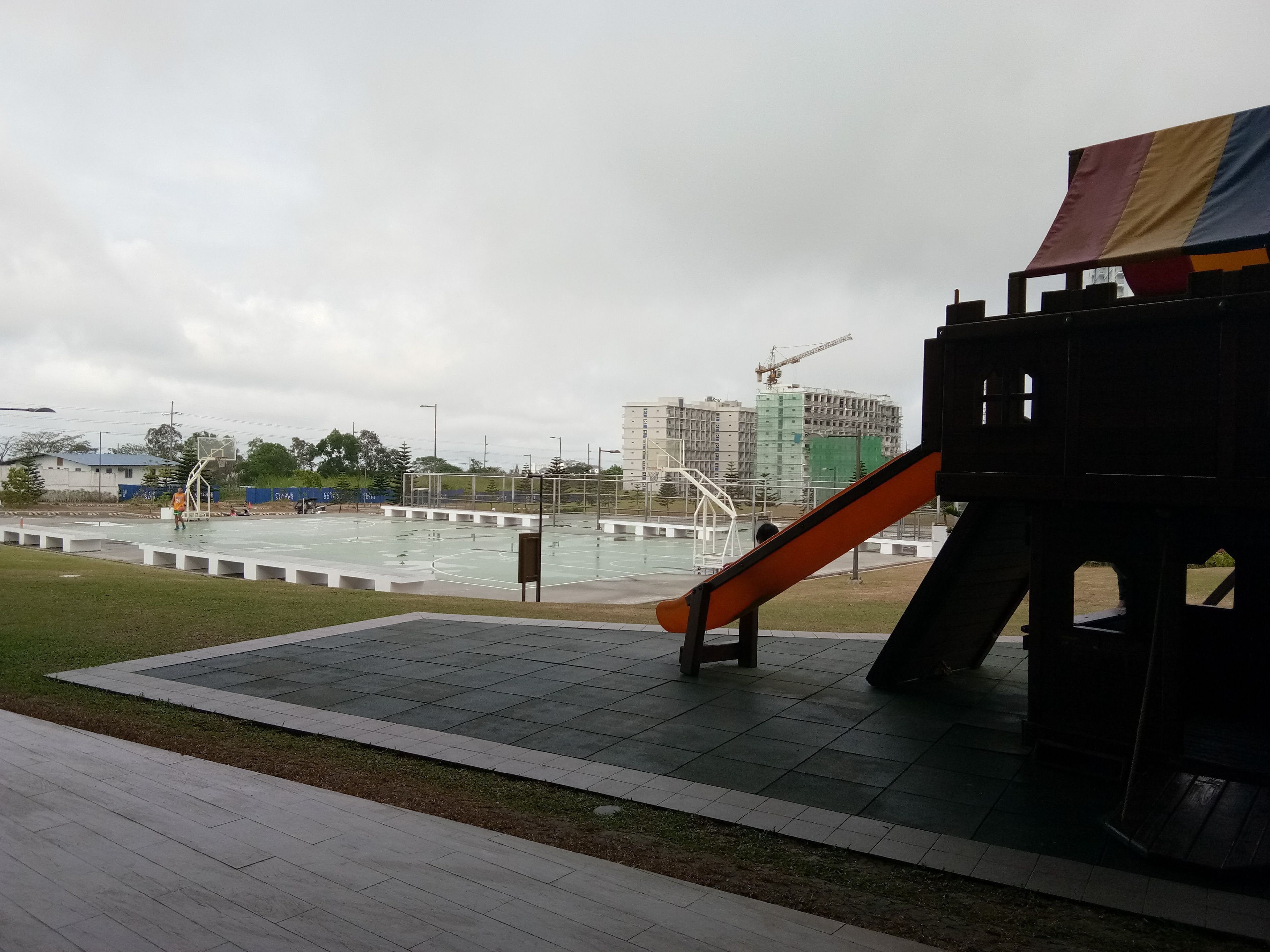 An outdoor basketball court with a slide, surrounded by a grassy area and a playground structure.