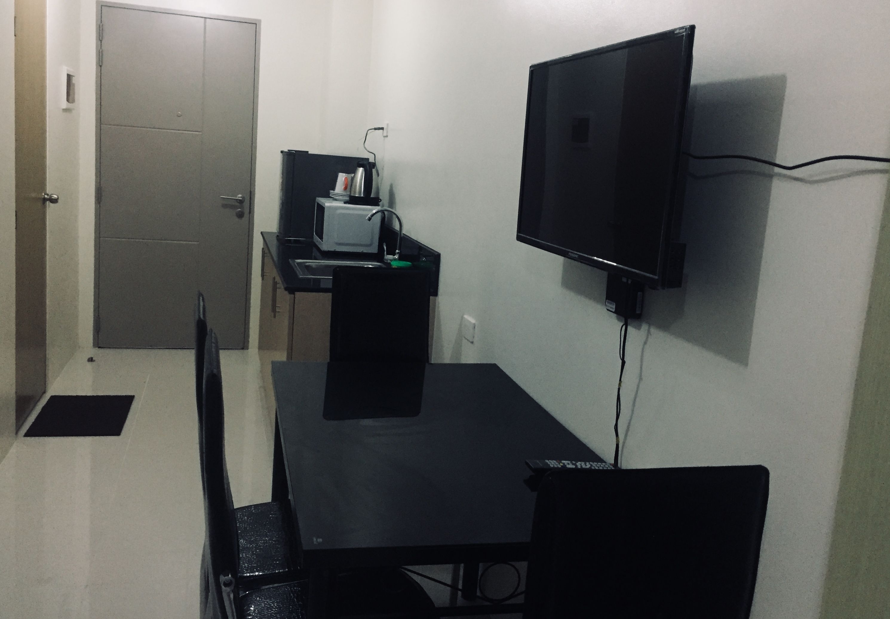 A small room with a black table, a chair, a TV mounted on the wall, a microwave on a shelf, and a door.