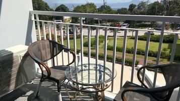 A balcony with two wicker chairs and a glass table, overlooking a street with parked cars and greenery.