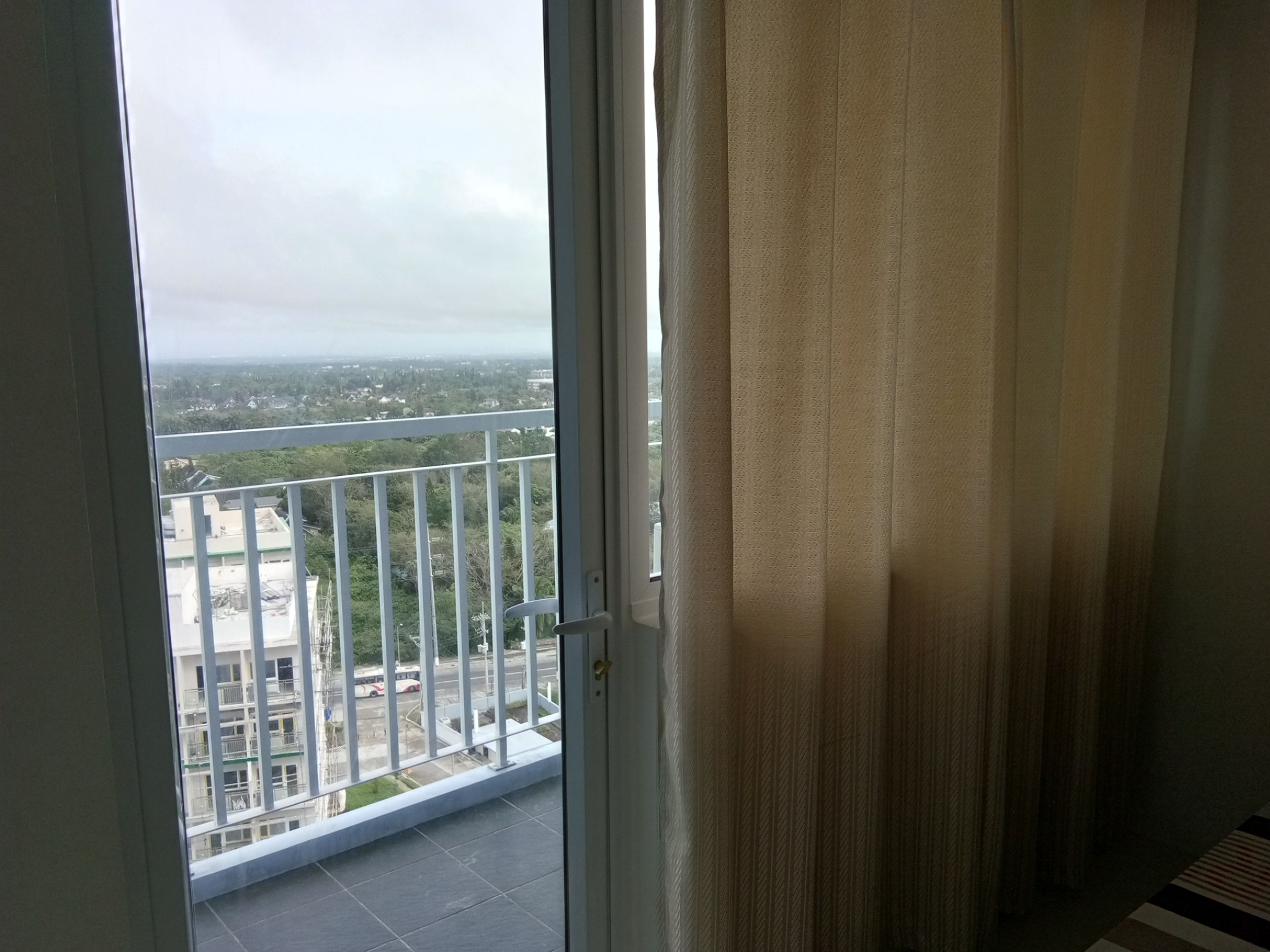 Un balcón con vistas a un paisaje urbano, barandilla blanca y una puerta con cortina.