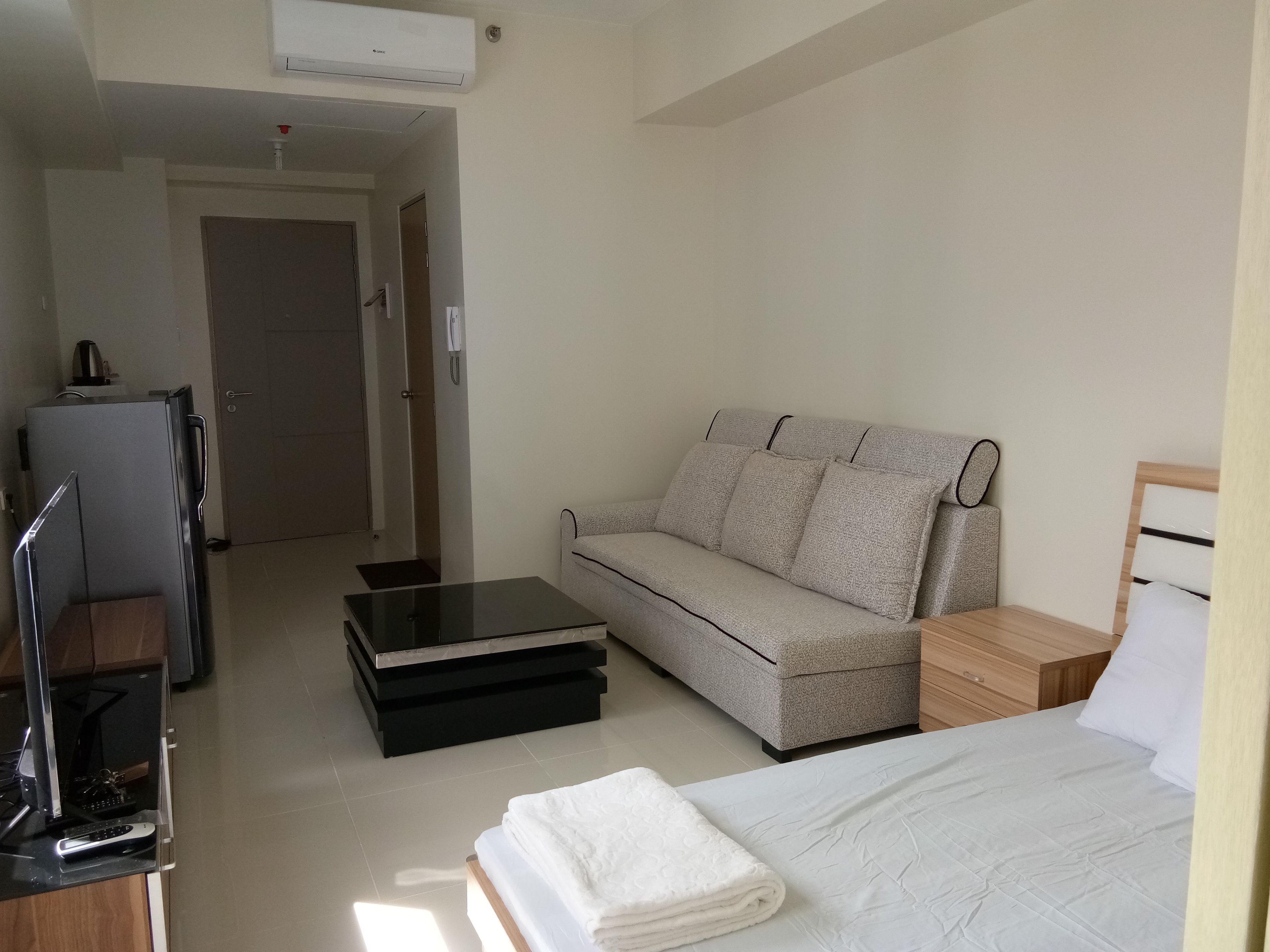 A compact hotel room with a bed, a sofa, a small table, a desk with a computer, and a refrigerator.