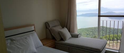 Ein Schlafzimmer mit einem Bett, einem grauen Sessel und einem großen Fenster mit Blick auf einen See und Berge.