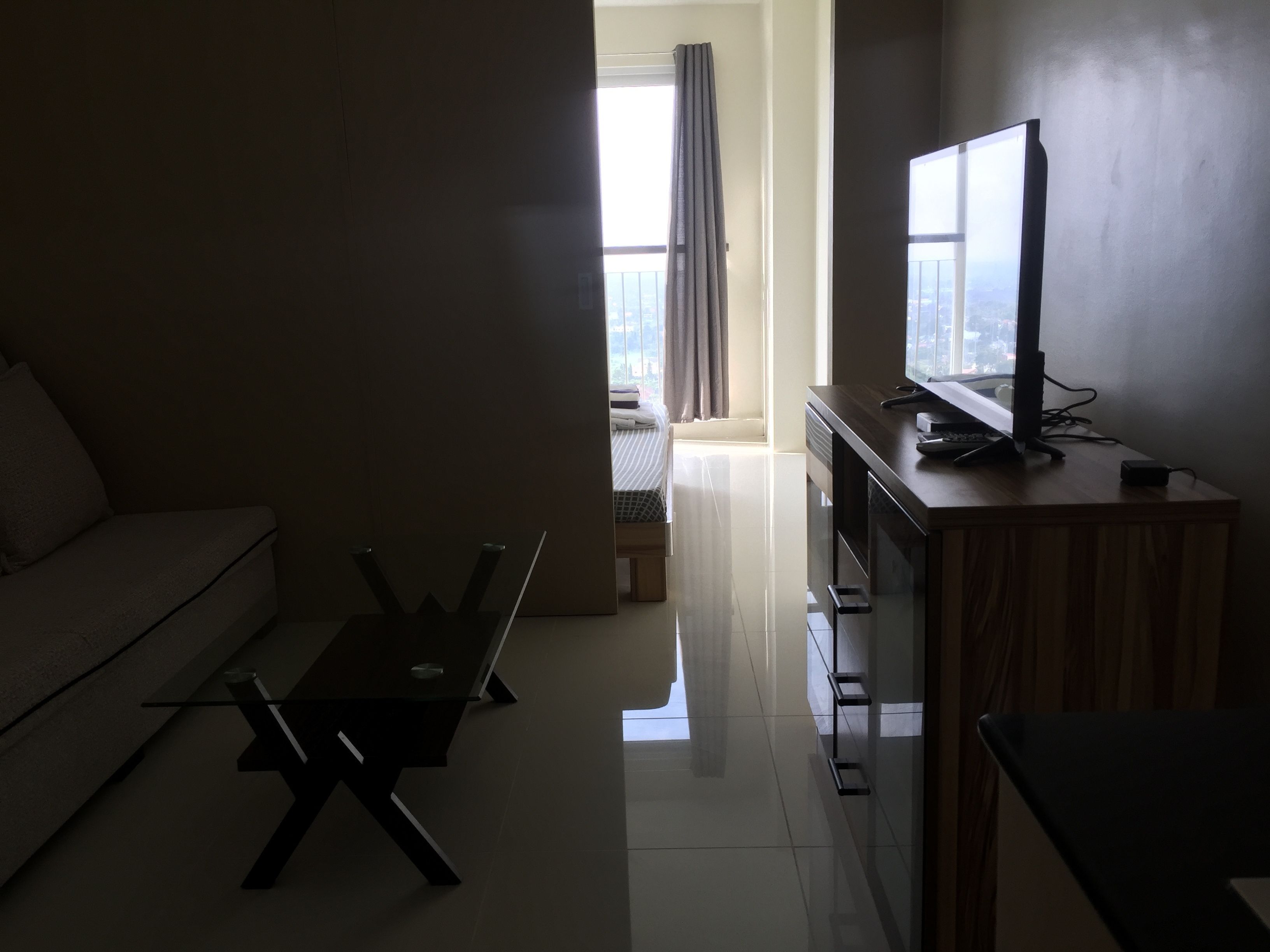 A modern living room with a sofa, a coffee table, a TV on a wooden stand, and a view of a balcony with curtains.
