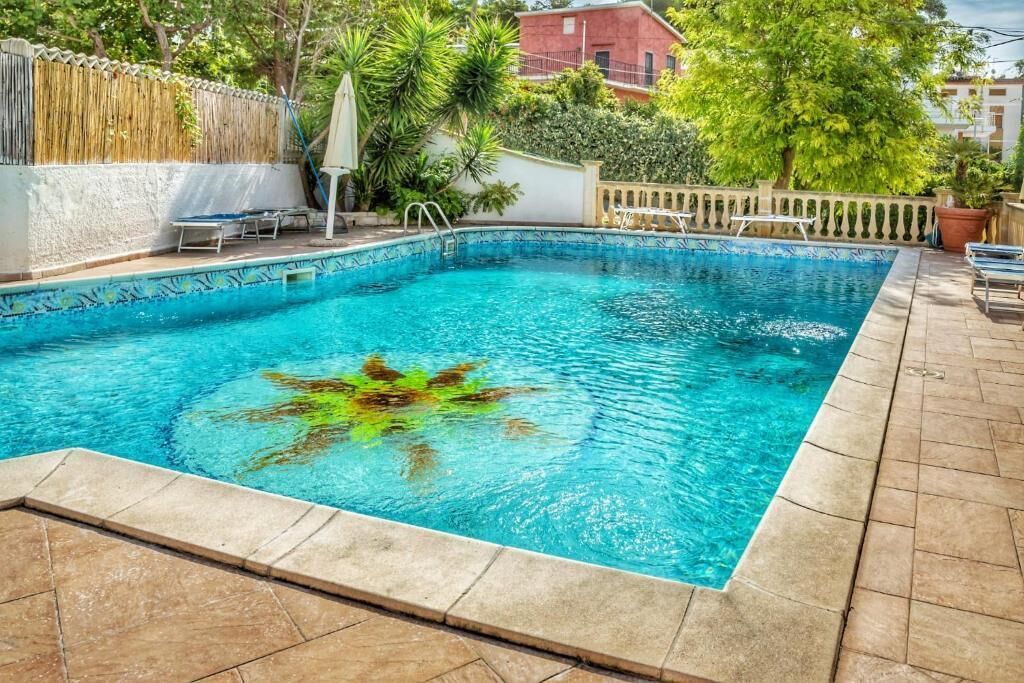 A rectangular swimming pool with a tiled border, surrounded by a patio area with chairs and a fence.