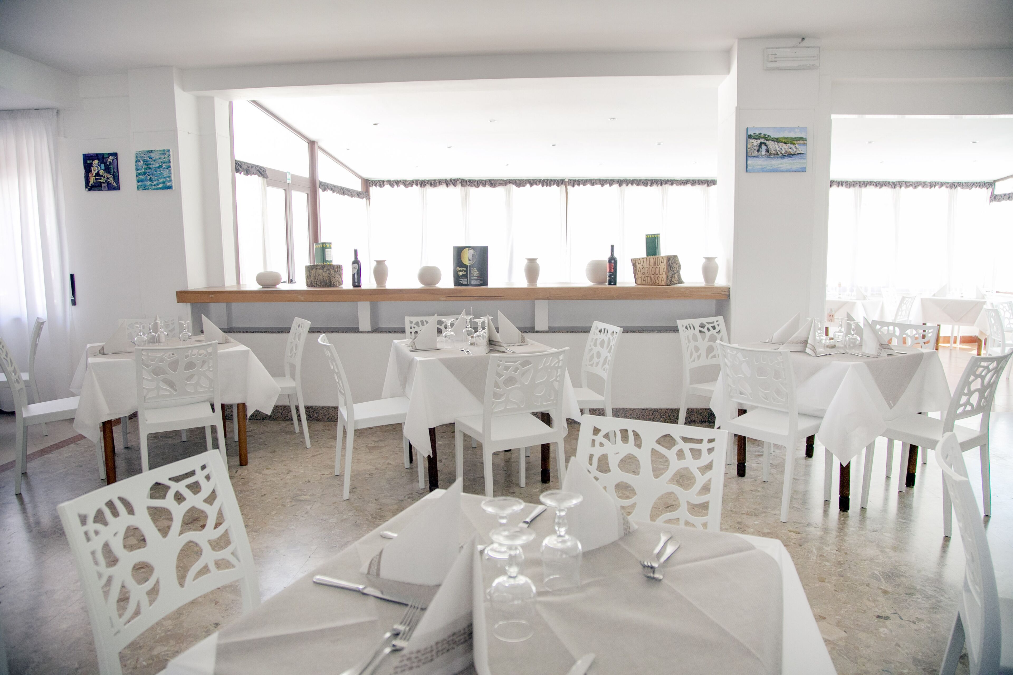 A dining area with white tables and chairs, a wooden shelf with decorative items, and a view through large windows.