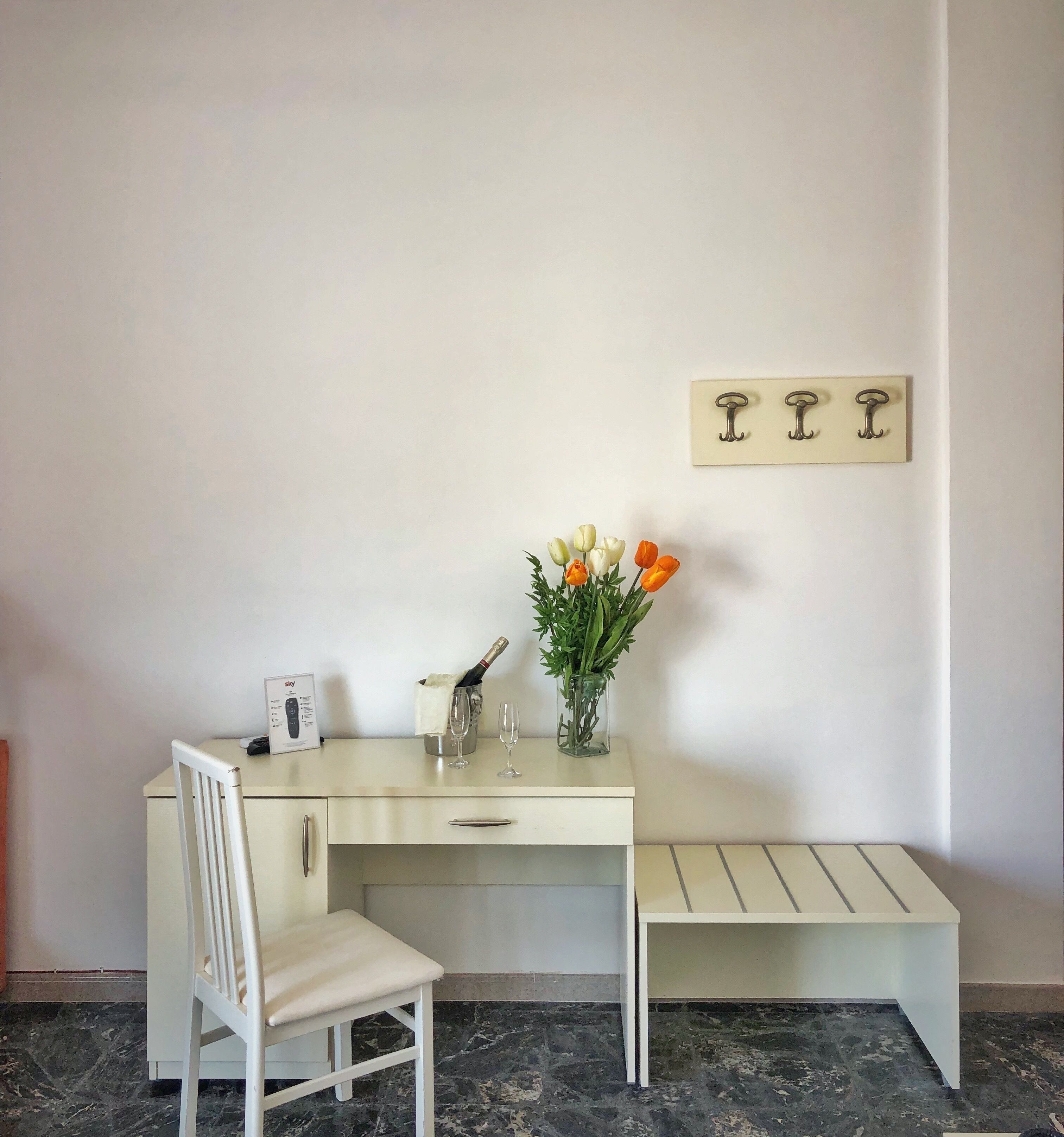 Un bureau blanc avec une chaise, un vase rempli de fleurs, une bouteille de champagne et une télécommande posés sur le bureau.
