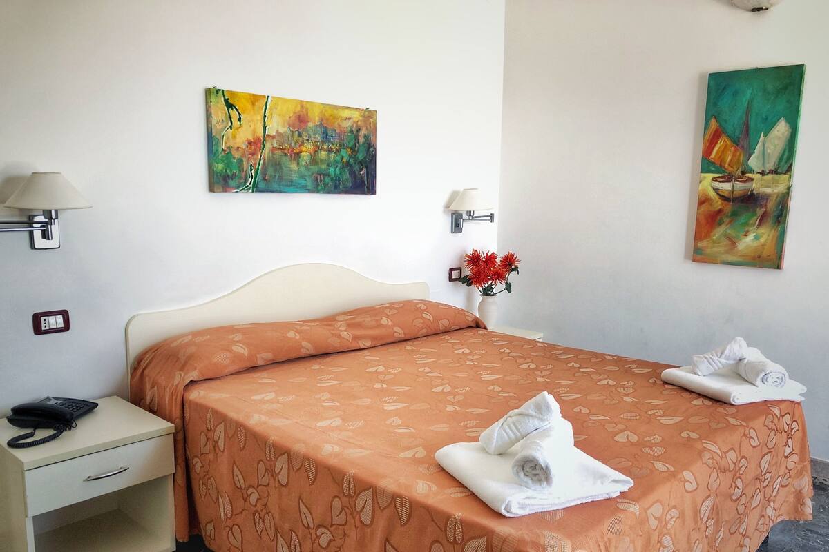 Ein Hotelzimmer mit Bett, Nachttisch, Lampe, einem Gemälde und einer Blumenvase.