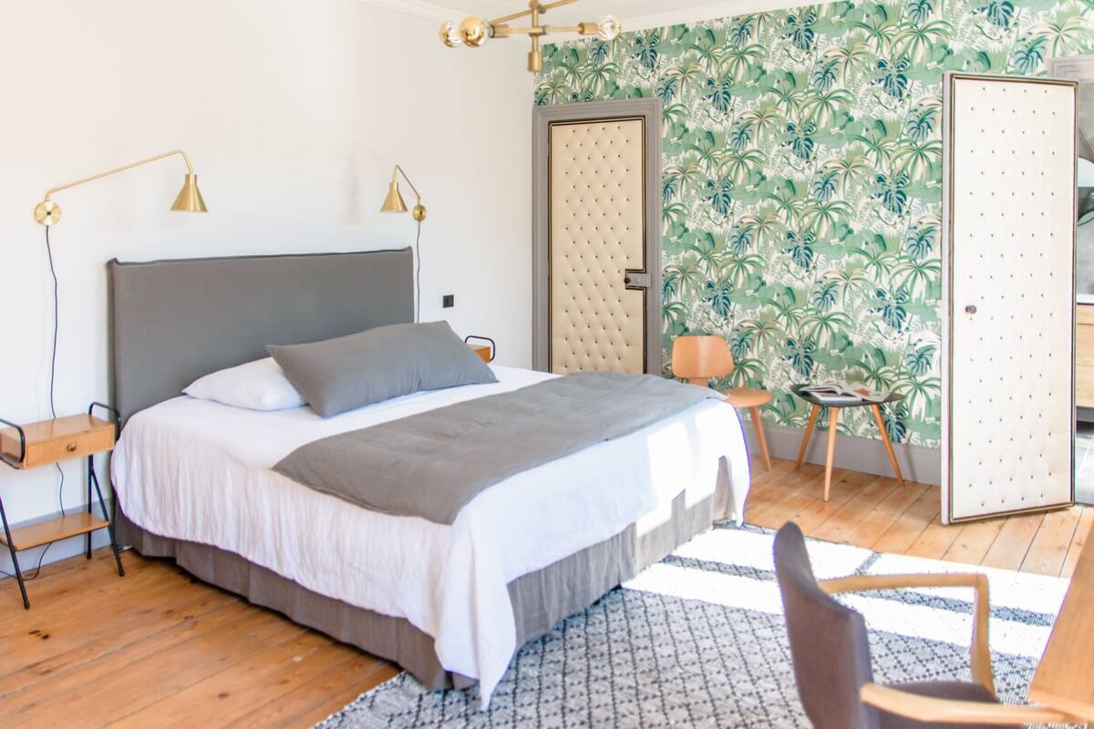 A neatly arranged bedroom with a large bed, a bedside table, a lamp, and a door with a patterned wallpaper.