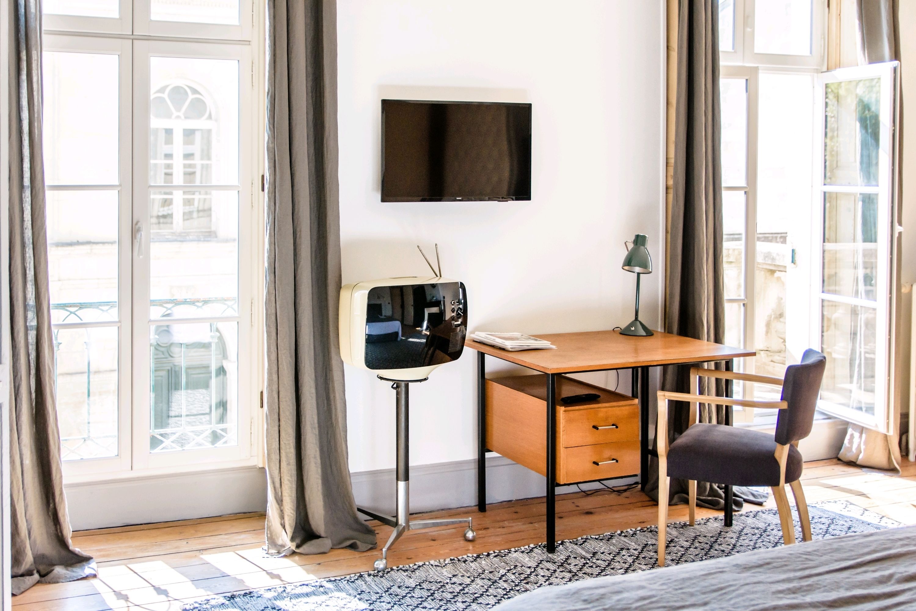 A room with a bed, a desk with a chair, a television on the wall, and large windows with curtains.