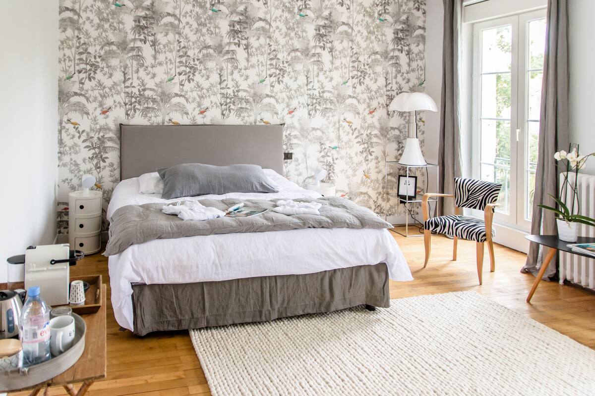 A bedroom with a large bed, a bedside table, a chair, a lamp, and a window with curtains.