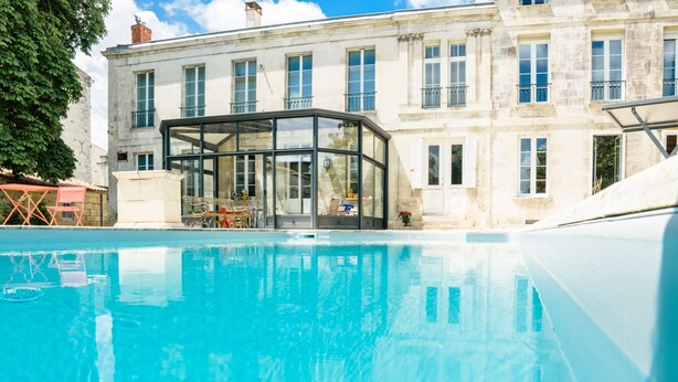 A swimming pool with a glass enclosure, a building with multiple windows and balconies, and a clear blue sky.
